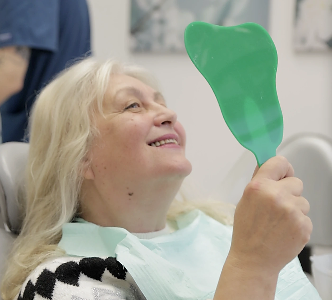 A smiling patient looks at her teeth in a hand mirror in the dental chair at LumiDent